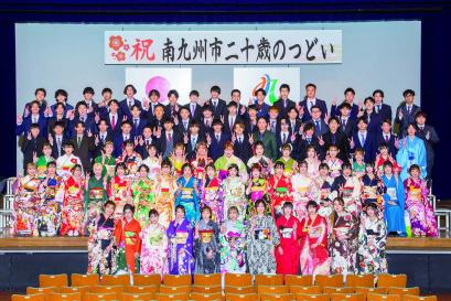 「祝南九州市二十歳のつどい」と書かれた横断幕が掲げられた舞台の上と下に、色とりどりの振袖を着た女性と、袴やスーツを着た男性の新成人が6列に並んで写した記念写真