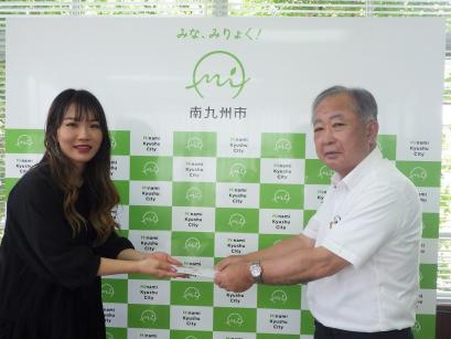 南九州市のバックボードの前で桐木平氏からの寄付を塗木市長が受け取っている写真