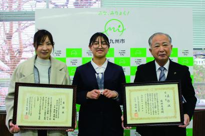 南九州市のバックボードの前で、受賞者の若林さんを挟んで左右に立った男女が額に入った表彰状をこちらへ見せて写した記念写真