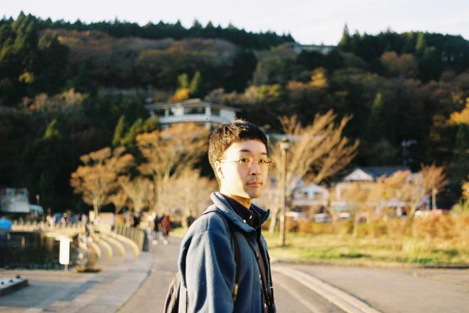奥に山が広がっている広場で、メガネをかけて身体を斜めにして立っている南九州市地域おこし協力隊・青木健太朗さんの写真
