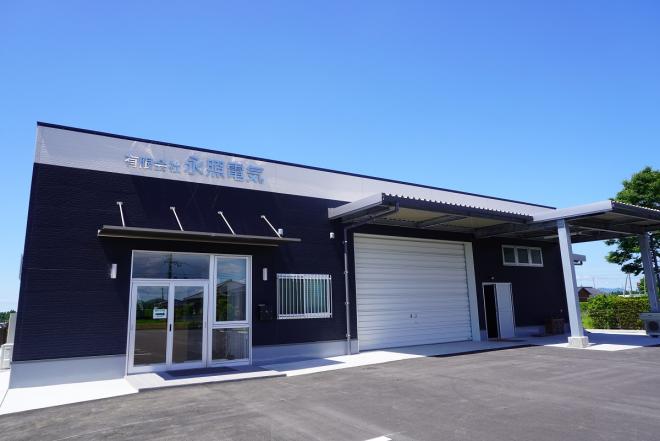 ネイビーを基調とした建物で入口の上に看板があり、右隣にシャッターが設置されている有限会社永照電気の写真