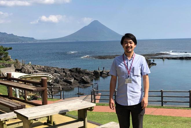海岸の向こう側の開聞岳が見える公園で、笑顔で立っているブルーのシャツを着た南九州市地域おこし協力隊・原本太郎さんの写真