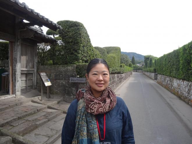 綺麗に剪定された植木が並んだ知覧武家屋敷群で、笑顔で首にスカーフを巻いて立っている南九州市地域おこし協力隊・岩崎泰依さんの写真