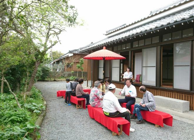 庭に赤い敷物が敷かれた長椅子と傘が設置されお茶会をしている方々にお茶を運んでいる南九州市地域おこし協力隊・川口塔子さんの写真