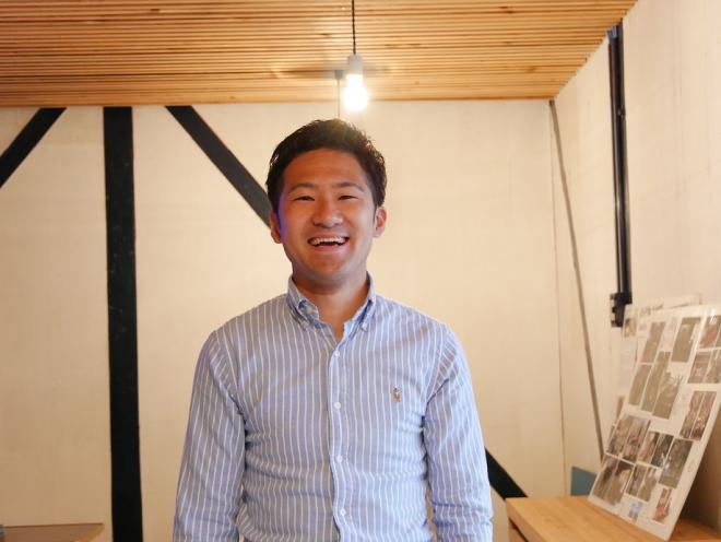 ボードにたくさんの写真が飾られた部屋で、笑顔で立っている南九州市地域おこし協力隊・蔵元恵佑さんの写真