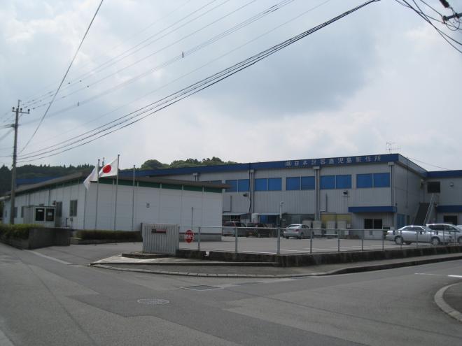 入口に国旗が掲げられ、青い屋根で2階建ての建物の日本計器鹿児島製作所(工場)の写真