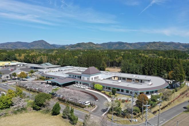 奥に山脈が広がり、横に長く楕円の形をした建物で、玄関前に駐車場がある音野舎を上空から撮影した写真