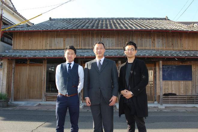 木造のモダンな日本家屋の前で笑顔で立ち、スーツを着た男性を挟み横1列に並んでいる(左)南九州市地域おこし協力隊の蔵元恵佑さん(右)前迫昇吾さんの写真