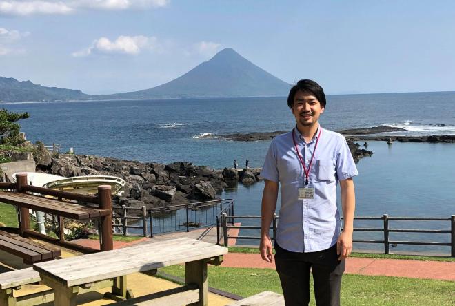 奥に開聞岳が見える公園で、ブルーのシャツを着た南九州市地域おこし協力隊・原本太郎さんが笑顔で立っている写真