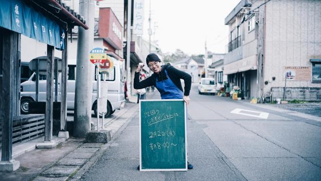 ネイビーの暖簾がかかったお店の前で黒板を持ち、エプロンを付けてガッツポーズを決めている南九州市地域おこし協力隊・福島花咲里さんの写真