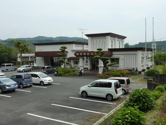 駐車場が隣接しており、屋根と柱の下部が一部がえんじ色で全体が白い外壁の知覧地区公民館の外観の写真