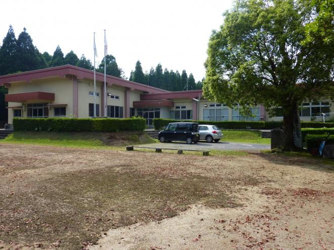 林の手前に建ち、周囲を低い生垣に囲まれた、白壁で柱や屋根が薄ピンク色のL字型平屋建ての松山地区公民館の建物外観の写真