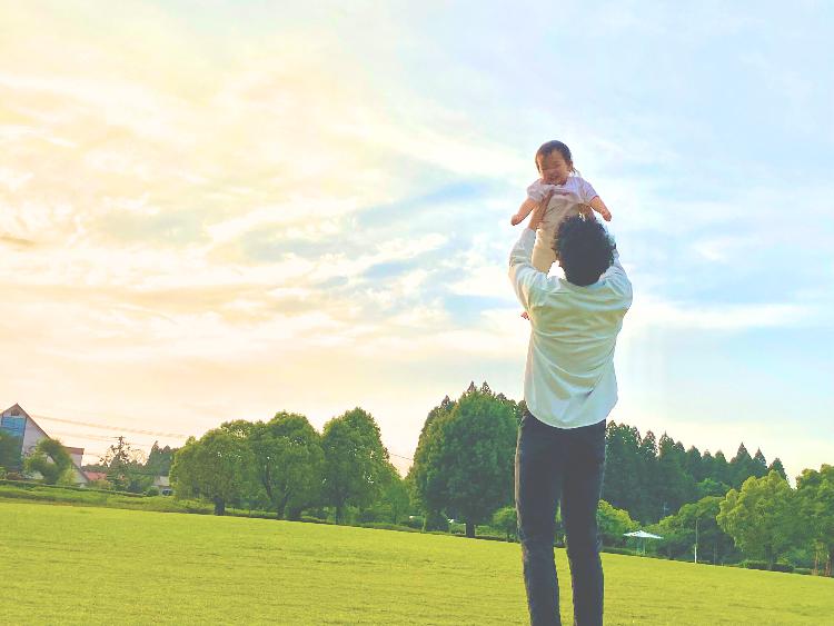 夕焼けの空を背景に、幼児の両脇を支えて高く持ち上げている男性の写真