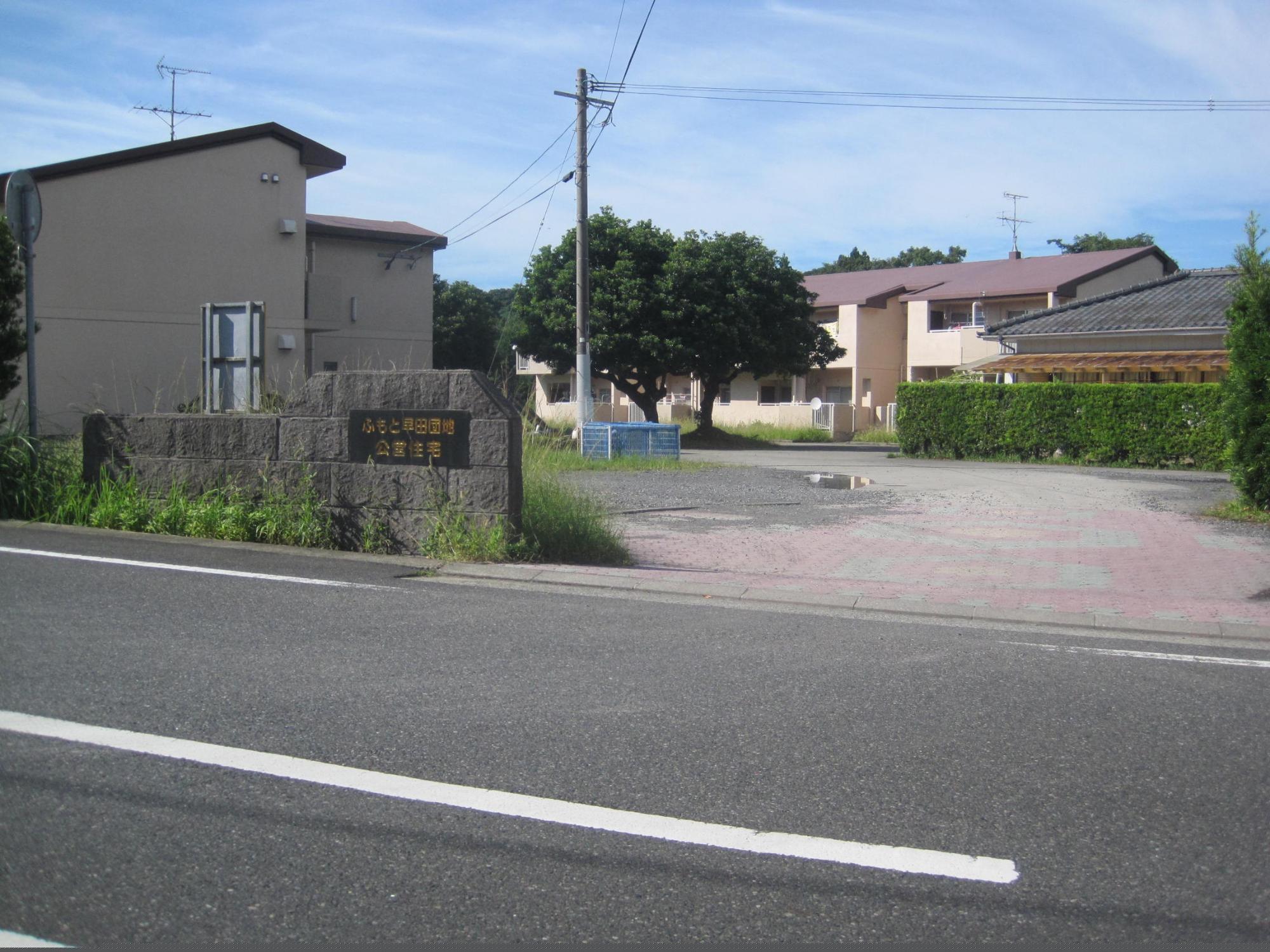 道路を挟んだ奥左側に門柱があり、その奥左側と右奥に2階建ての市営住宅が建っている公営・特公賃ふもと早田団地の写真