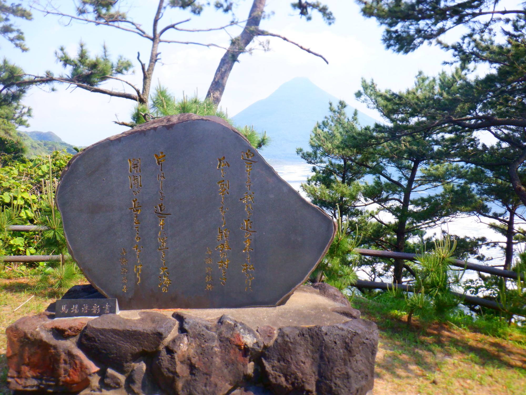 歌碑が設置されている場所の後方に開聞岳が木々の間から見えている写真