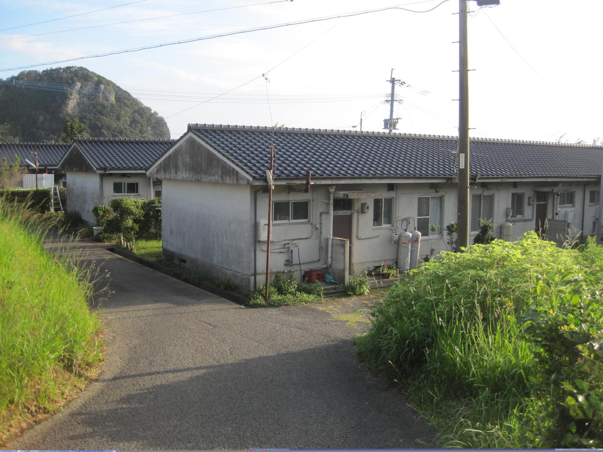 両脇に草が生い茂っている道路の先に建っている横長で1階建ての市営住宅 公営ふもと坂上団地2棟の写真