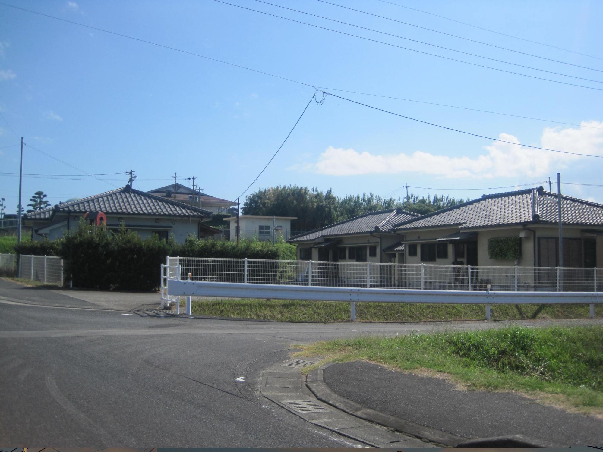 ガードレールの後ろに白いフェンスがあり、フェンスの奥に瓦屋根1階建て住宅が並んでいる市営住宅 公営大川団地の写真