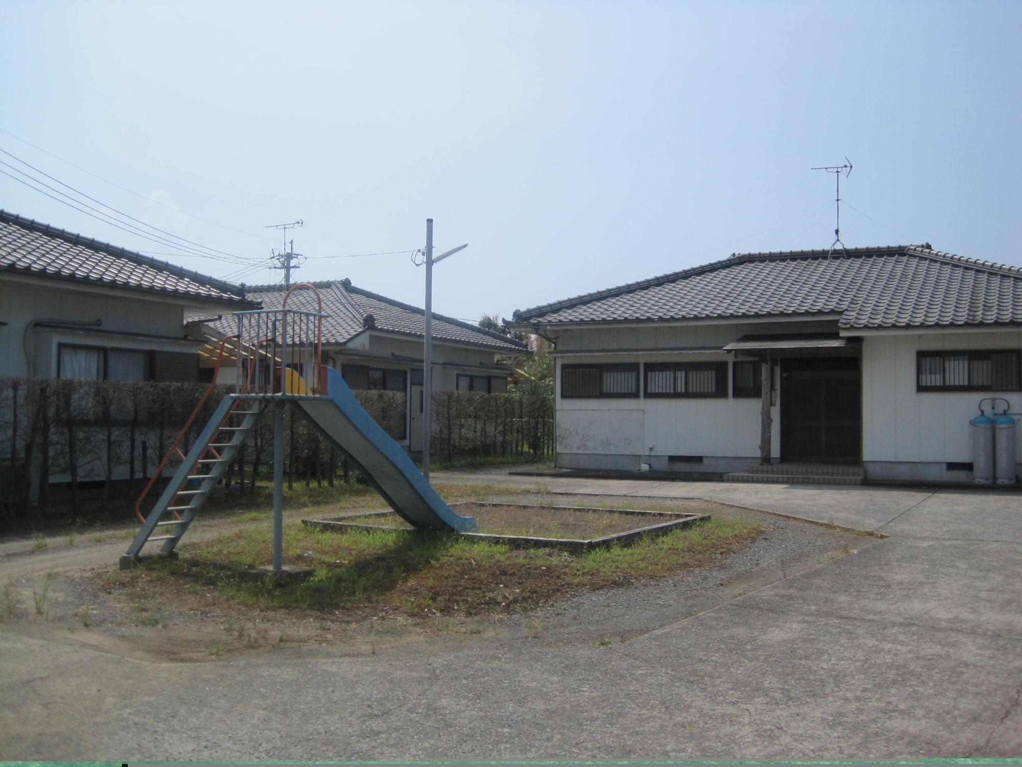青い滑り台がある広場の奥と左側の垣根の奥に、瓦屋根1階建て住宅が3軒建っている市営住宅 特公賃大川団地の写真
