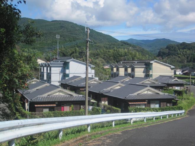 下り坂左側の土地が低くなっている場所に瓦屋根木造平屋建ての住宅が約10軒、奥に3階建ての住宅2棟が建っている市営住宅 公営山田比良団地（木平・鉄筋）の写真