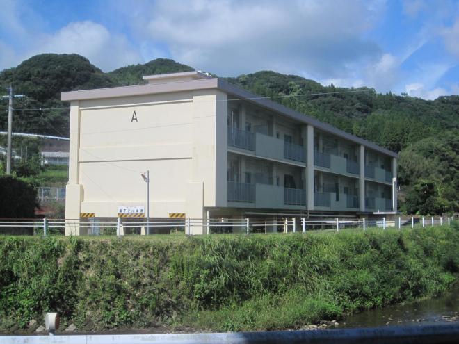 手前に川、奥に山がある場所に3階建てで団地の側面に「A」と書かれている市営住宅 公営鼻面田団地A棟の写真