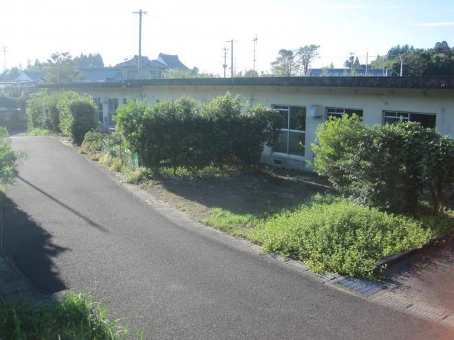 舗装された通路の右側に人の背丈ほどの木々が植えられており、その奥に屋根が平らで横長の1階建ての建物が建っている市営住宅 公営浮辺団地の写真