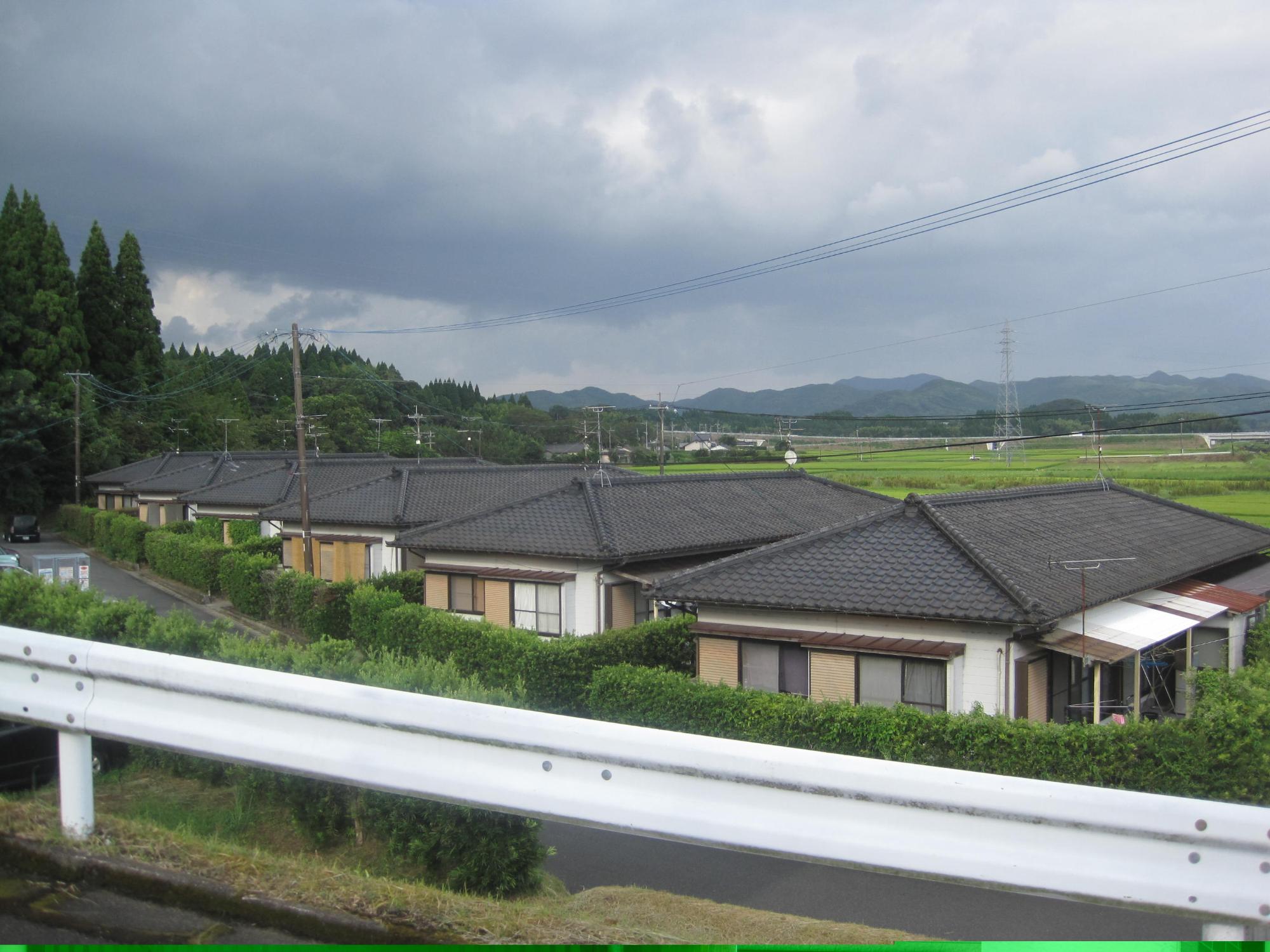 ガードレール下の田園風景が広がる手前に、瓦屋根の1階建て住宅が6軒並んでいる市営住宅 公営瀬世団地の写真