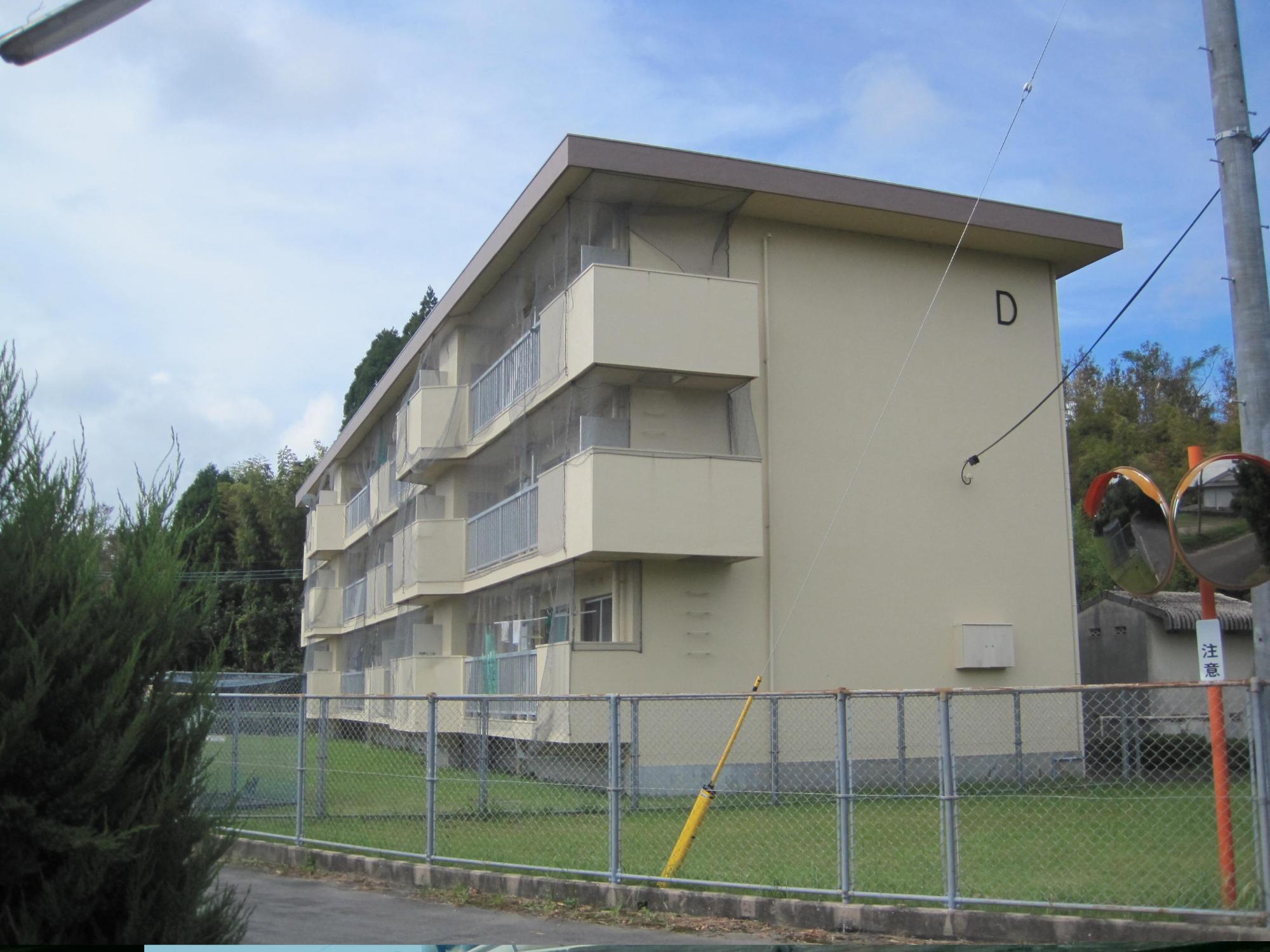 金網のフェンスの奥に全体がクリーム色で外壁右上の角に「D」とある3階建ての市営住宅 公営瀬世団地D棟の写真