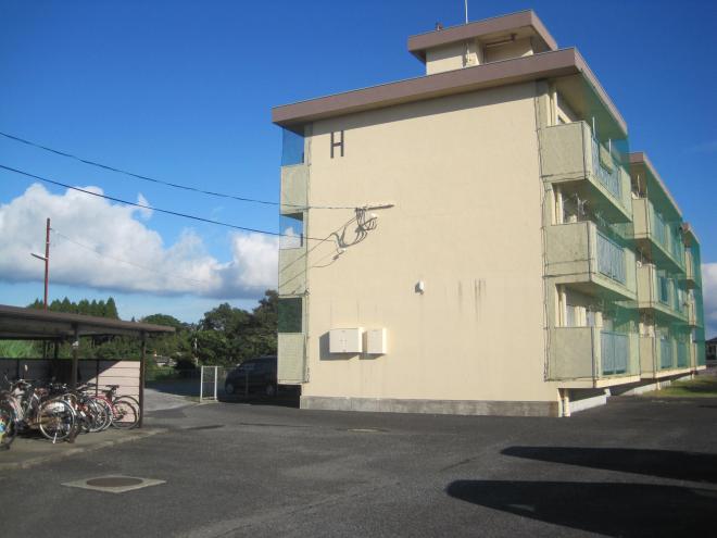 手前左側に屋根付きの自転車置き場、建物側面左上の角に「H」とあり、ベランダに鳥除けのネットがされている市営住宅 公営霜出団地H棟の写真