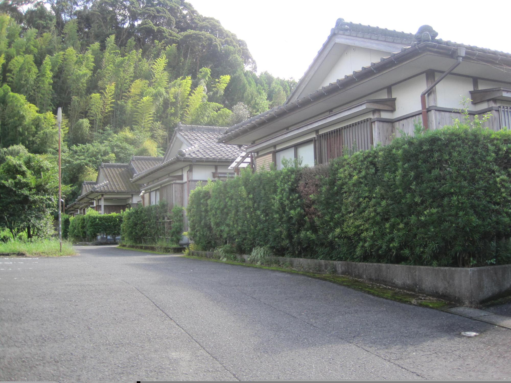 奥に雑木林がある手前に、垣根で囲まれた瓦屋根1階建ての住宅が3軒並んでいる市営住宅 特公賃本町団地の写真