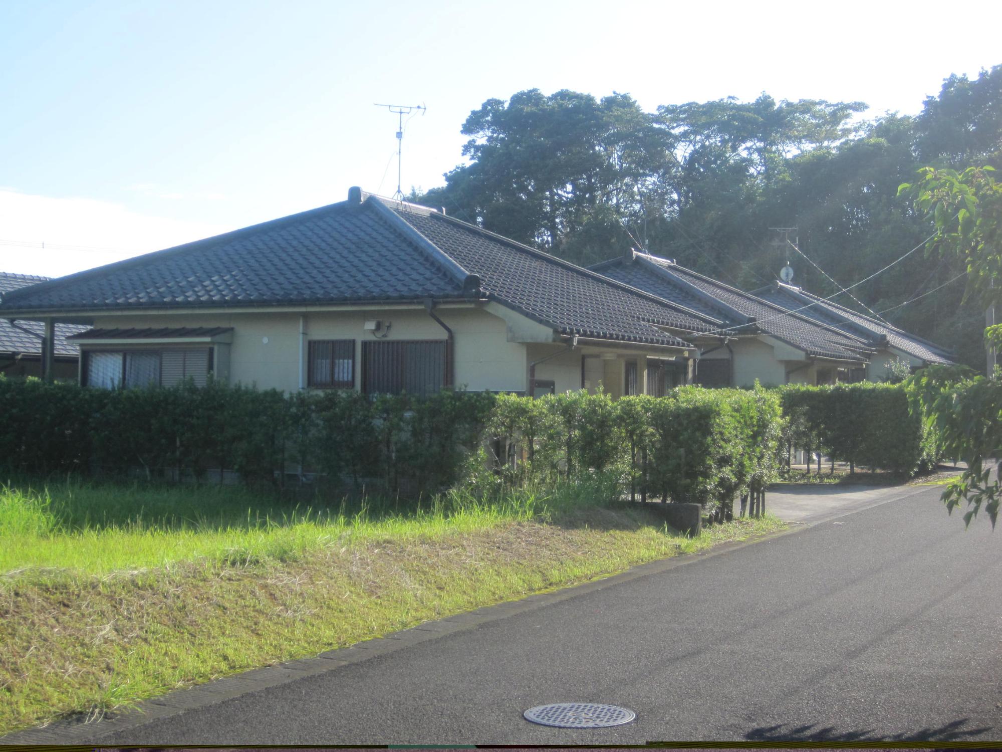 左側の空き地の横に垣根で囲まれている瓦屋根平屋建て住宅が3軒並んで建っている市営住宅 特公賃みずほ団地の写真