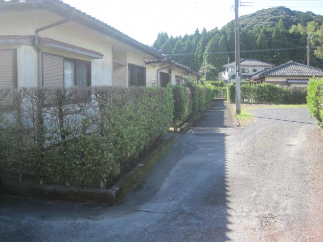 砂利道がある左側に建っている、垣根で囲まれ横に長い平屋建ての市営住宅 一般永里団地の写真
