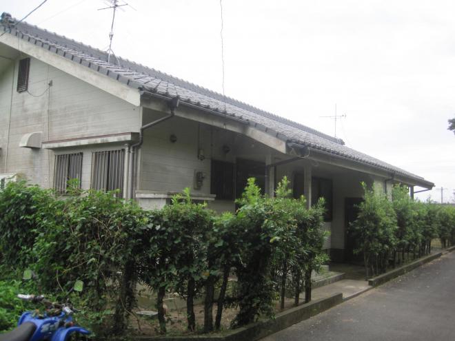垣根の奥に建っている三角屋根平屋建ての市営住宅 一般丸尾団地を斜め前から写した写真