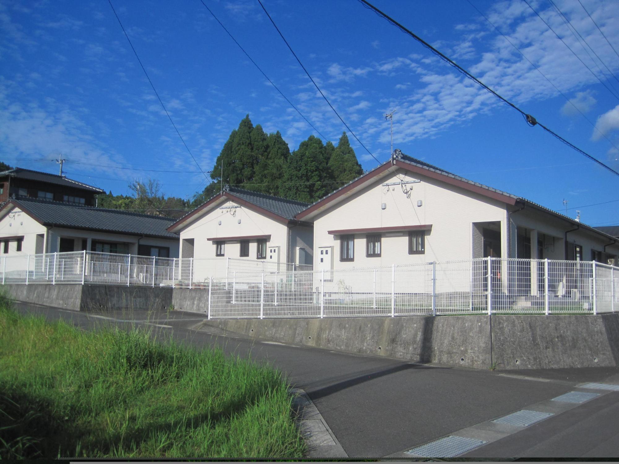 道路に面し、白い格子状のフェンスの奥に、三角屋根白い外壁の平屋建て住宅が3軒並んでいる市営住宅 公営緑ヶ丘団地の写真
