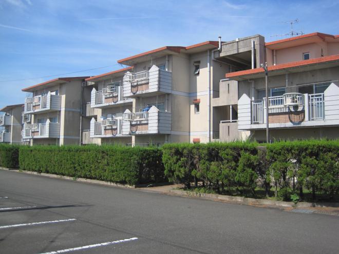 手前から駐車場、垣根、奥に屋根部分がオレンジ色の3階建てベランダ付きの建物が建っている市営住宅 公営・特公賃川辺松山団地の写真