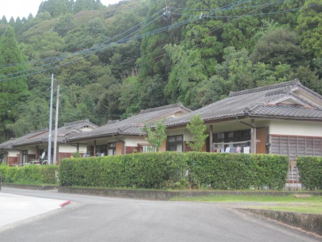 すぐ後ろに山がある手前に建っている、垣根で囲まれた瓦屋根平屋建ての住宅が3軒ほど並んでいる市営住宅 公営・一般本門団地の写真