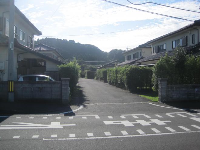 「とまれ」と書かれている道路の奥にブロック塀でできた入り口があり、入り口から真っ直ぐ伸びた道路の両側に垣根で区切られている2階建ての住宅が6軒ほど並んでいる市営住宅 公営高田団地の写真