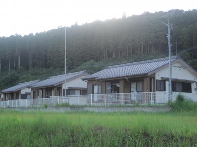 手前に原っぱ、奥に雑木林が見える場所に建っている三角屋根平屋建ての住宅が3軒並んだ市営住宅 公営両添団地の写真