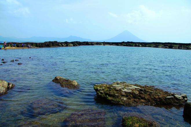 浅瀬の海岸が広がる奥に開聞岳がうっすらと見えている番所公園の写真