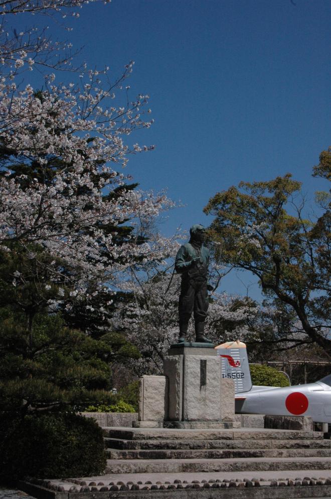 桜の花が咲き、澄み切った青空の下に建っている特攻隊員の銅像と、シルバーの機体に赤い日の丸が描かれた飛行機が設置されている写真。