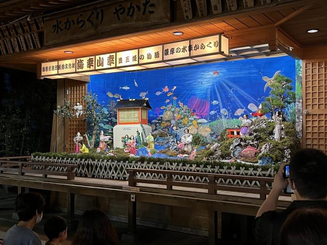 様々な魚が泳ぐ海中を背景とし、複数のからくり人形が置かれている舞台の写真
