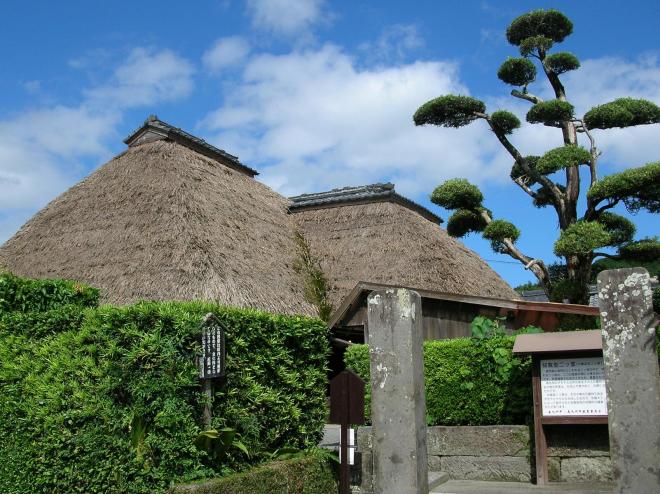 生垣の上から傾斜のついた茅葺屋根が見える知覧型二ツ家民家の外観写真