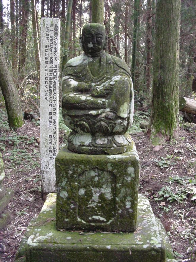 杉林の中、にこやかな笑顔を浮かべる仏像が設置された大野氏四代の供養塔がある勝目城跡と白い案内看板