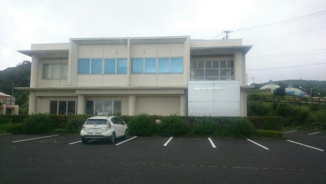 白壁で2階建ての建物が、駐車場と生垣の先に建っている南九州市頴娃特産品開発研究センターの外観写真