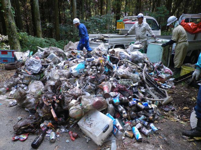 山の中で3人のヘルメットとを被った男の人が缶や燃えないゴミなど大量のごみを集め、軽トラック2台に乗せようとしている写真