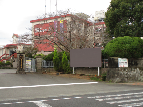 レンガ色と白を基調とした奥に体育館、手前に3階建ての校舎があり門の前は横断歩道がある川辺高等学校の写真