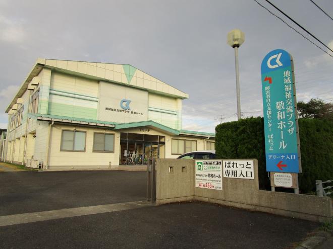 右側の塀の側に地域福祉交流プラザ敬和ホールと書いた薄い緑の看板があり、クリーム色を基調とした2回建ての地域地域福祉交流プラザ敬和ホール外観の写真