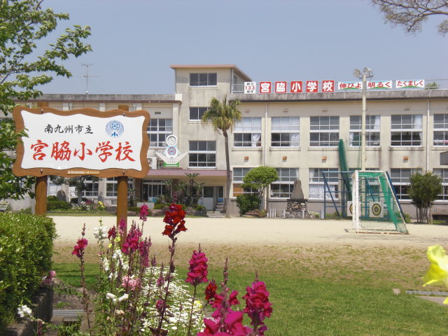 赤、白、ピンク色の綺麗な花が咲いている花壇の側に「南九州市立宮脇小学校」の立て看板が設定され、奥に広い運動場と2階建ての校舎が見える宮脇小学校の外観写真