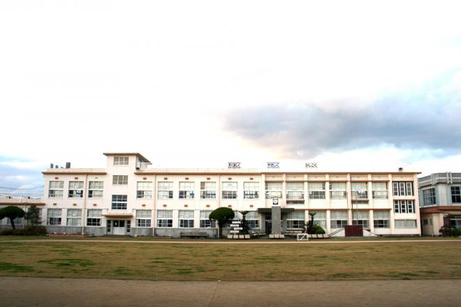 屋上の柵に「やさしく、かしこく、たくましく」と書かれた横断幕が掛けられ、白壁で2階建ての南九州市立粟ヶ窪小学校を校庭から撮影した写真