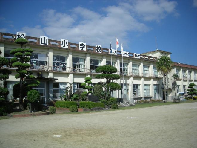 周囲に様々な形をした手入れされた植木が植栽され、屋上の柵に「松山小学校 かしこく、やさしく、元気よく」と書いた看板がつけられた、白壁で2階建ての南九州市立松山小学校の建物外観の写真
