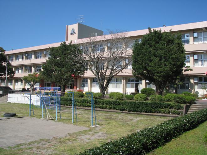 周囲に植木や大きな木が等間隔で植栽されている3階建ての南九州市立知覧中学校の校舎を、鉄棒が設置されている校庭側から撮影した写真